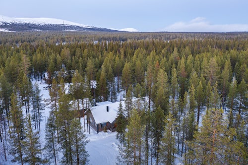 Drone Photography by UK London Freelance Drone Photographer Aerial of wooden cabin in the remote forest with snow covered woods and trees landscape and mountains in Lapland Finland Europe